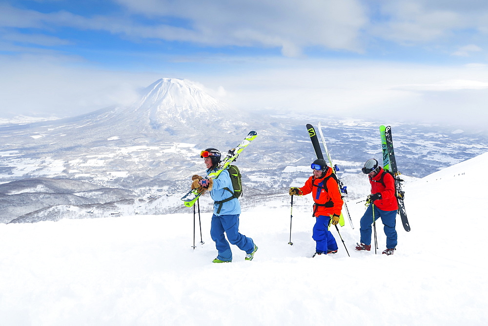 With the volcano Yotei in the background a female and two male backcountry skiers are hiking to the summit of mount Annupuri, in the ski resort Niseko United on the Japanese island of Hokkaido. Niseko United is comprised of four resorts on the one mountain, Annupuri (1,308m). 100km south of Sapporo, Niseko Annupuri is a part of the Niseko-Shakotan-Otaru Kaigan Quasi-National Park and is the most eastern park of the Niseko Volcanic Group. Hokkaido, the north island of Japan, is geographically ideally located in the path of consistent weather systems that bring the cold air across the Sea of Japan from Siberia. This results in many of the resorts being absolutely dumped with powder that is renowned for being incredibly dry. Some of the Hokkaido ski resorts receive an amazing average of 14-18 meters of snowfall annually. Niseko is the powder capital of the world and as such is the most popular international ski destination in Japan. It offers an unforgettable experience for all levels of skier and snowboarder. Mount Yotei in the background is often referred to as the "Mount Fuji of Hokkaido".