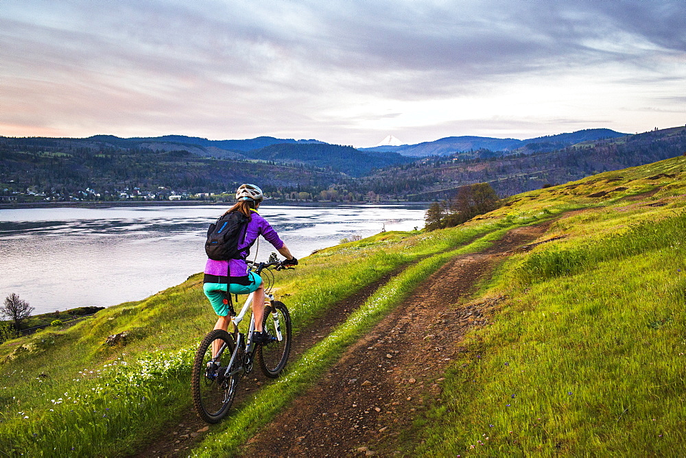 A young woman rides a mountain bike up a single-track trail through an open meadow under early morning sky with river in distance.