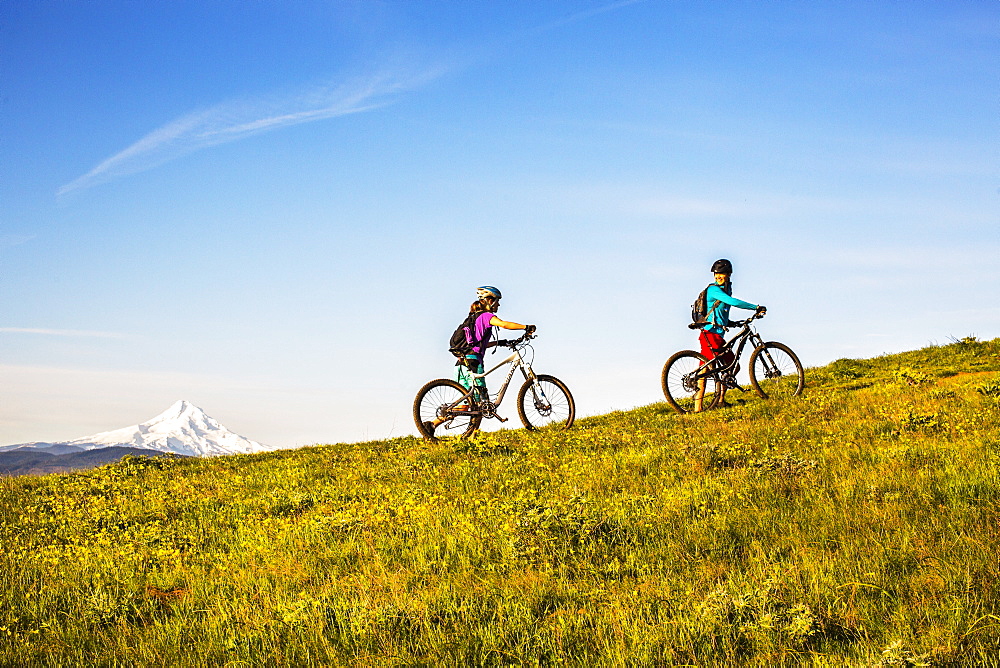 Two young women push mountain bikes up a single-track trail through an open meadow.