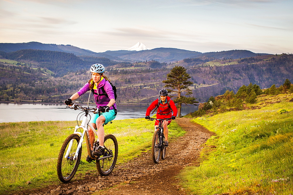 Two young women ride mountain bikes up a single-track trail with volcano in distance.