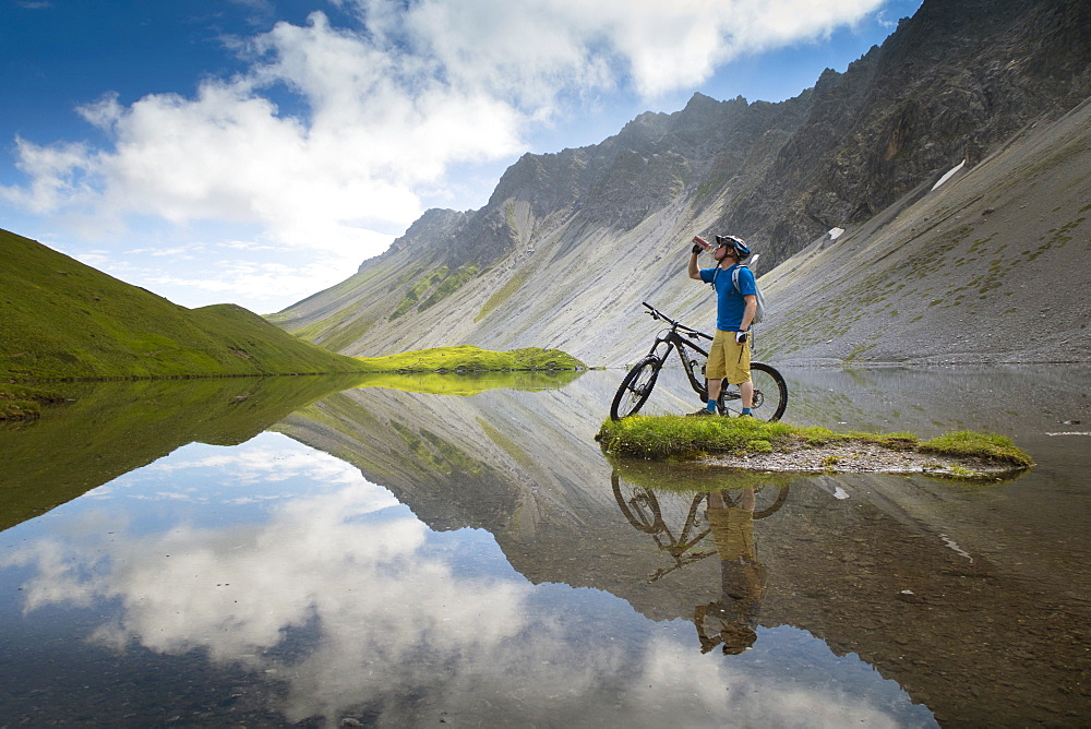 A Mountain Biker Is Drinking Water While Standing At A Beautiful Mountain Lake