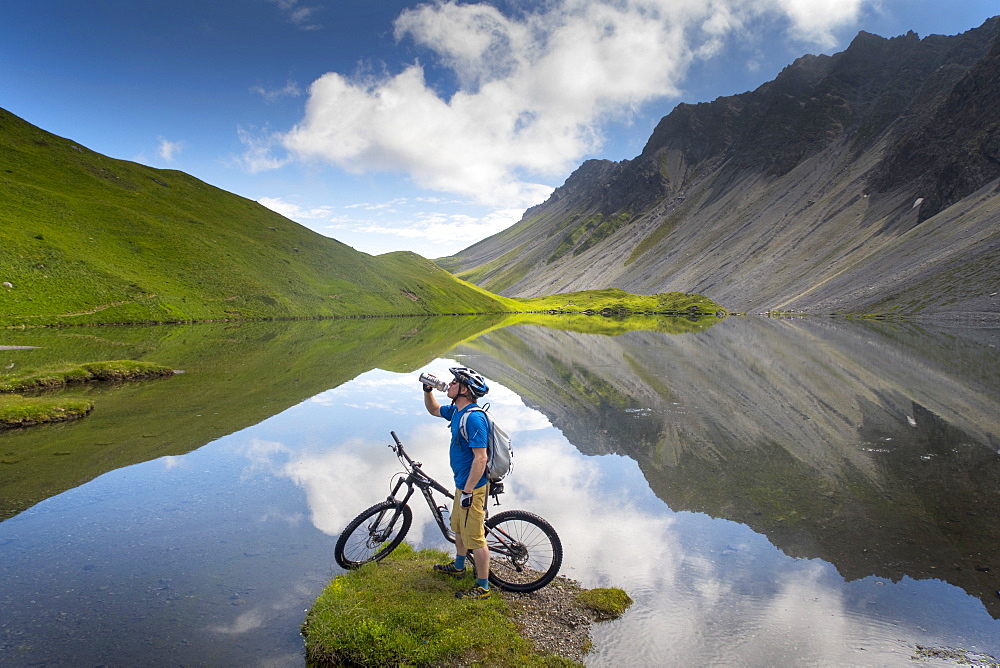 A Mountain Biker Is Drinking Water While Standing At A Beautiful Mountain Lake