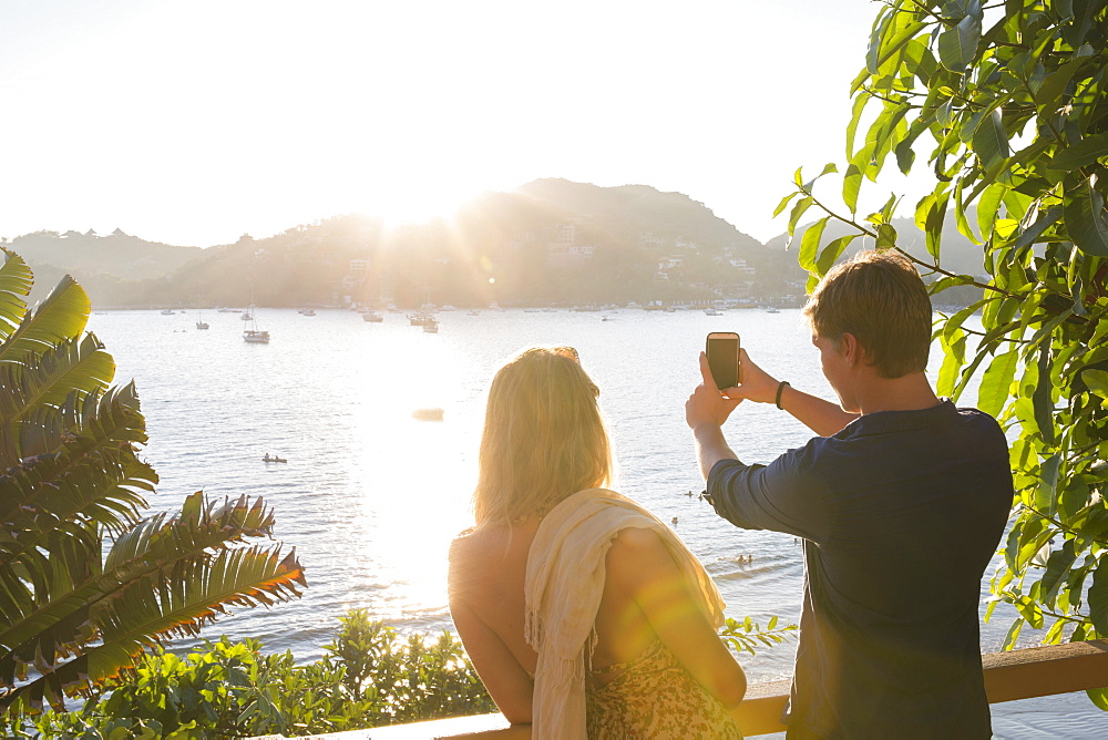 Young adult couple taking cell phone photo of harbor at sunset