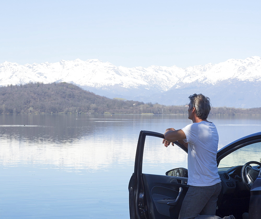 Mature man relaxes on door of car by lake