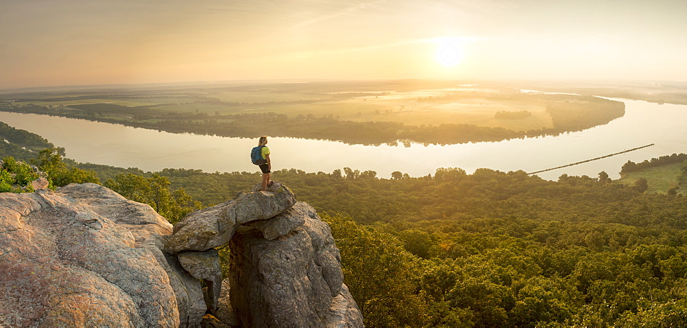 Woman standing on sandstone overhang watching sunrise from summit of Petit Jean Mountain above Arkansas River Valley 