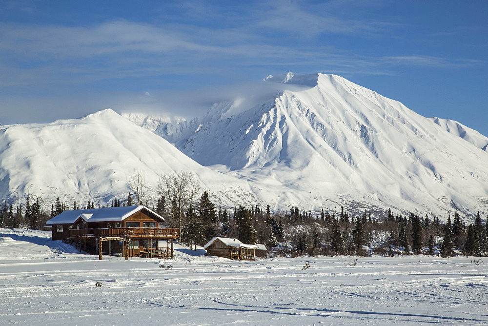 Log cabins in Alaska sit at the base of a frozen lake with snowy mountains in the background.