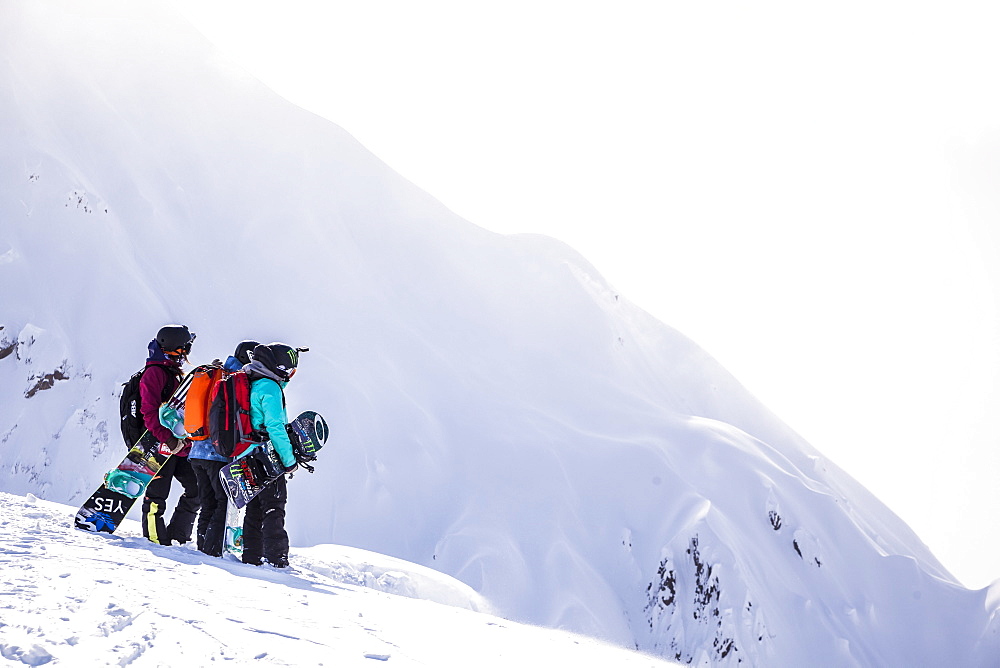 Professional snowboarders Robin Van Gyn, Helen Schettini, and Jamie Anderson, stand on a ridge and look down at a line they are about to ride on a sunny day in Haines, Alaska.