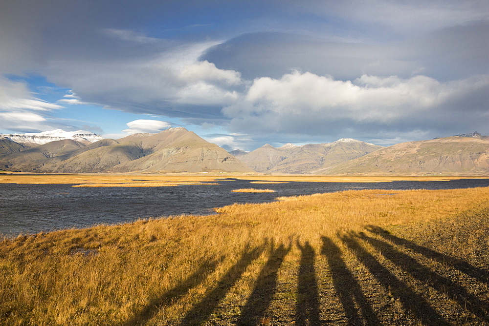 Long shadows of persons raising their hands projected on yellow grass with mountains and clouds. Photographed in golden light before sunset.