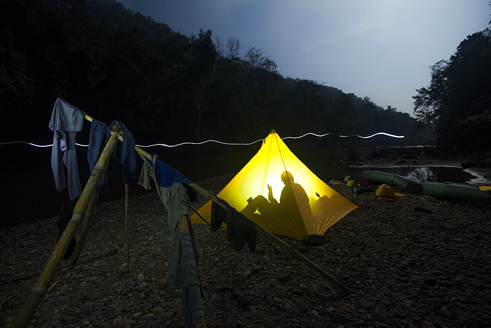 Robert Hahn reads in his tent as Ethan Welty walks past with his headlamp on at their camp on the Nam Ou River in Phou Den Din National Protected Area, Laos. The wooden tripod was built by hunters and fishermen who regularly camp and poach wild game within park boundaries despite (or perhaps without knowledge) that this is technically illegal.