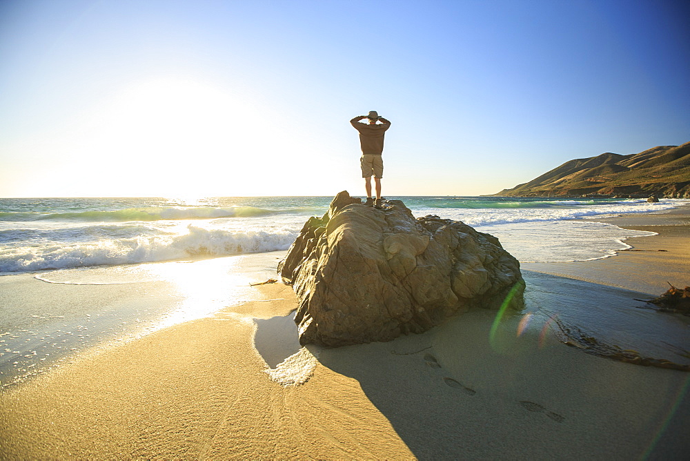 A man standing on a boulder at a beach looks out at the Pacific Ocean at sunset near Big Sur California.