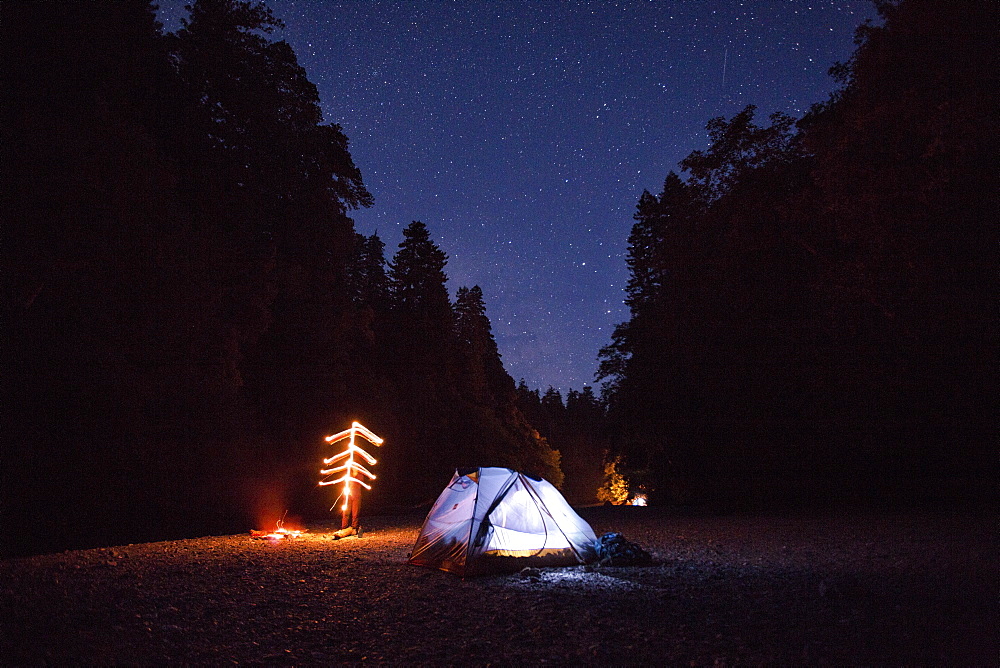 An illuminated tent sits next to a campfire on a river bed under a clear night sky with stars.