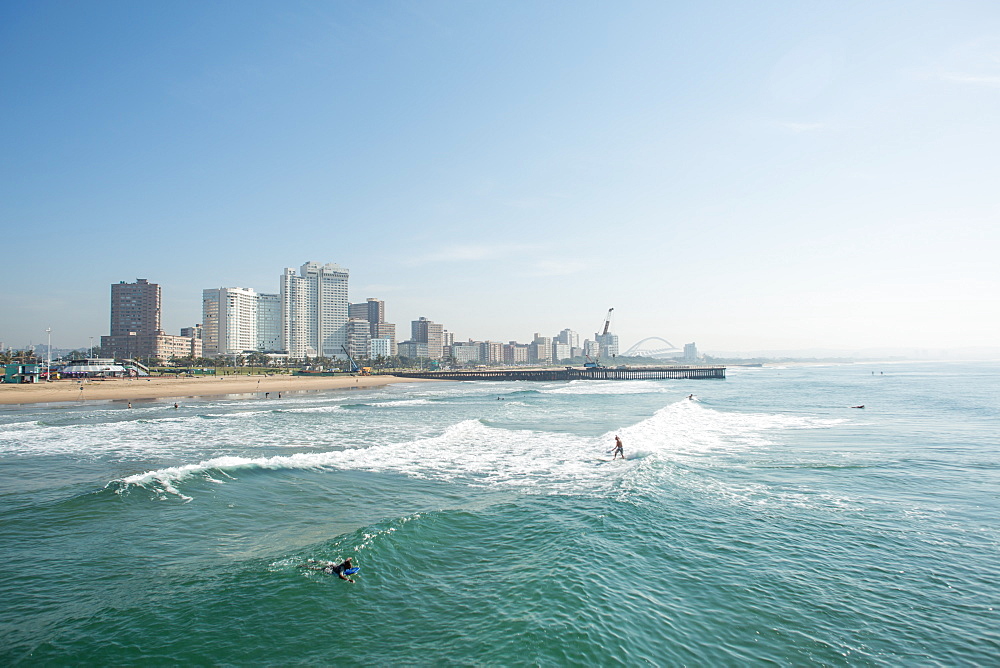 Surfers riding waves near pier on Golden Mile with skyline of Durban, South Africa