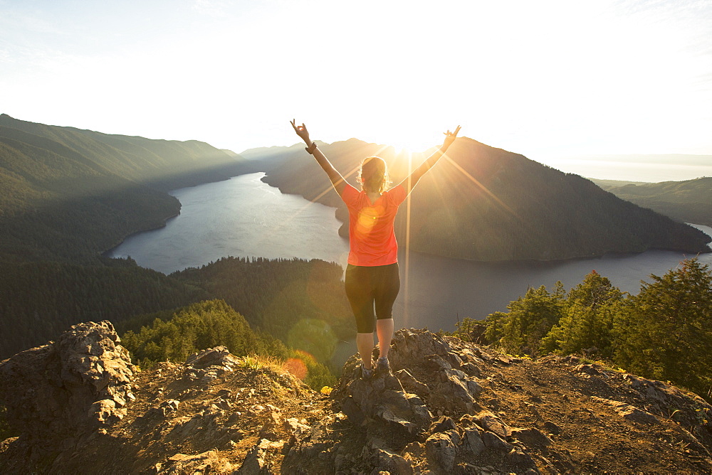 A female hiker in a red shirt celebrates reaching the top of the Mount Storm King trail in Olympic National Park at sunset.