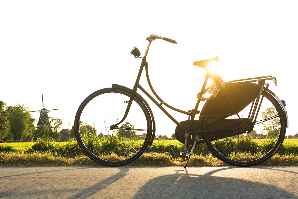 A vintage, worn traditionally Dutch bicycle propped up on the side of a bikepath, in the Dutch countryside at sunset, with a windmill in the background, in the province of Groningen, The Netherlands.