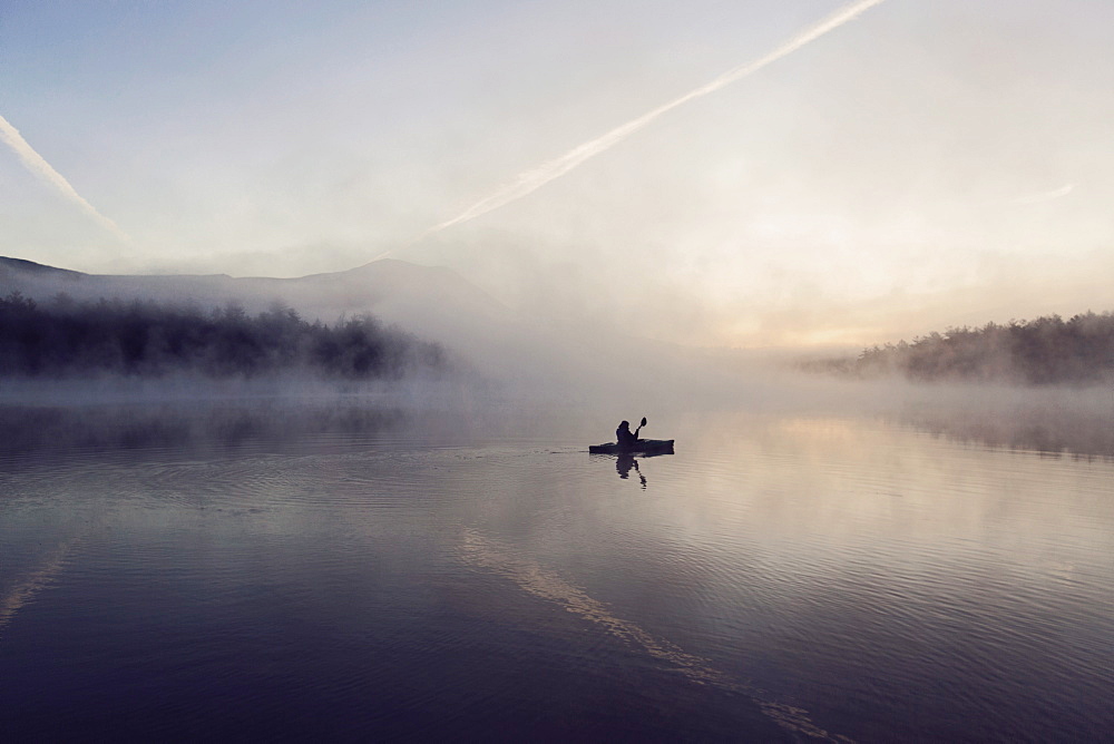 Young woman enjoys early morning paddle in kayak through mist on Daicey Pond in Maine's Baxter State Park, USA