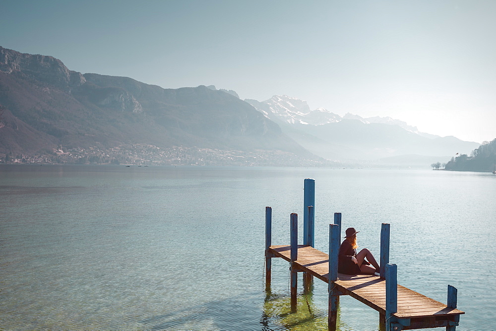 Woman sitting on jetty on lakeshore under clear sky with mountains in background, Annecy, Haute-Savoie,