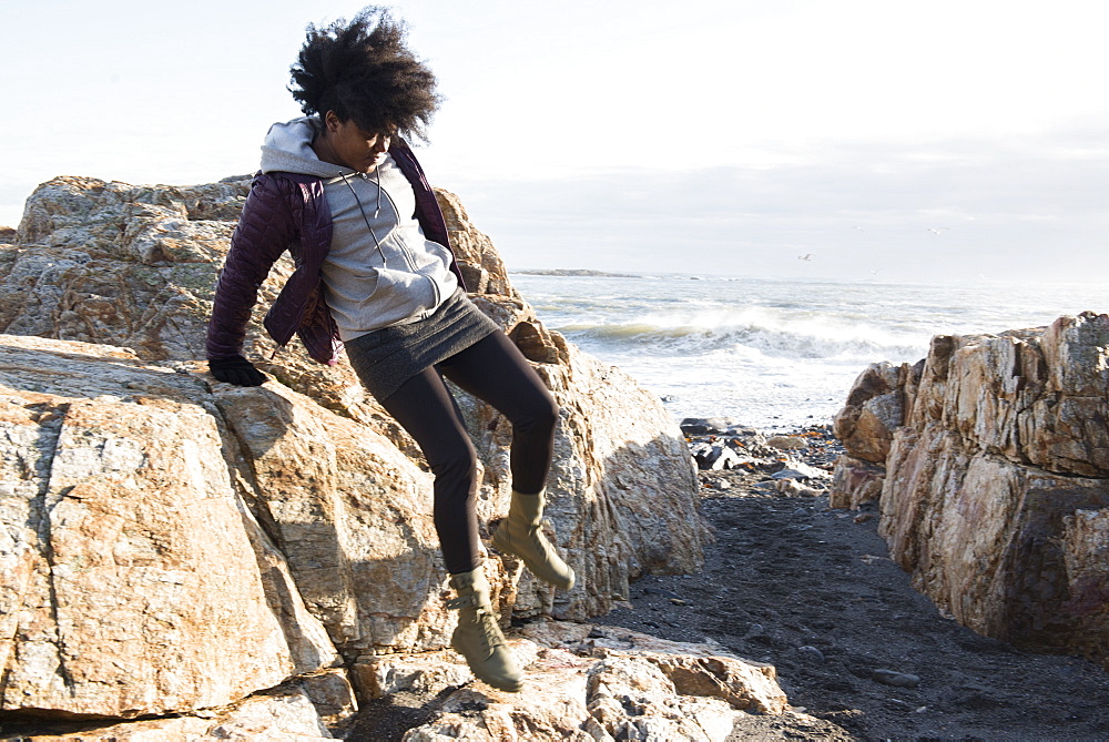 An African American woman in a purple jacket jumping down from a rock near the sea