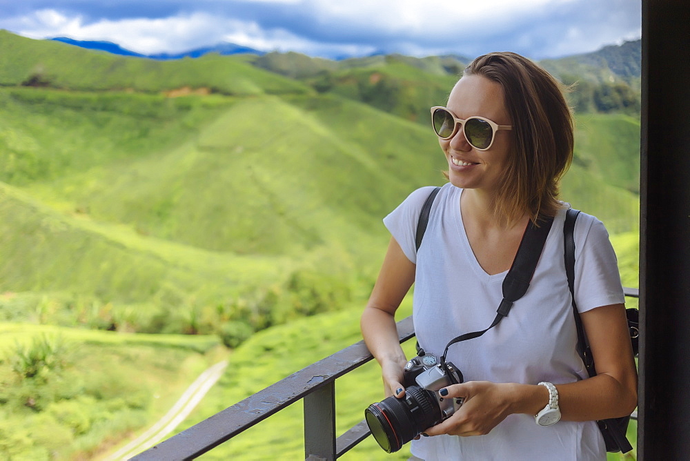 Waist up shot of smiling female tourist with sunglasses and camera at tea plantation, Cameron Highlands, Malaysia