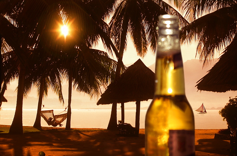 Beer bottle against beach with palm trees and woman in hammock at sunset, Badian, Cebu, Philippines