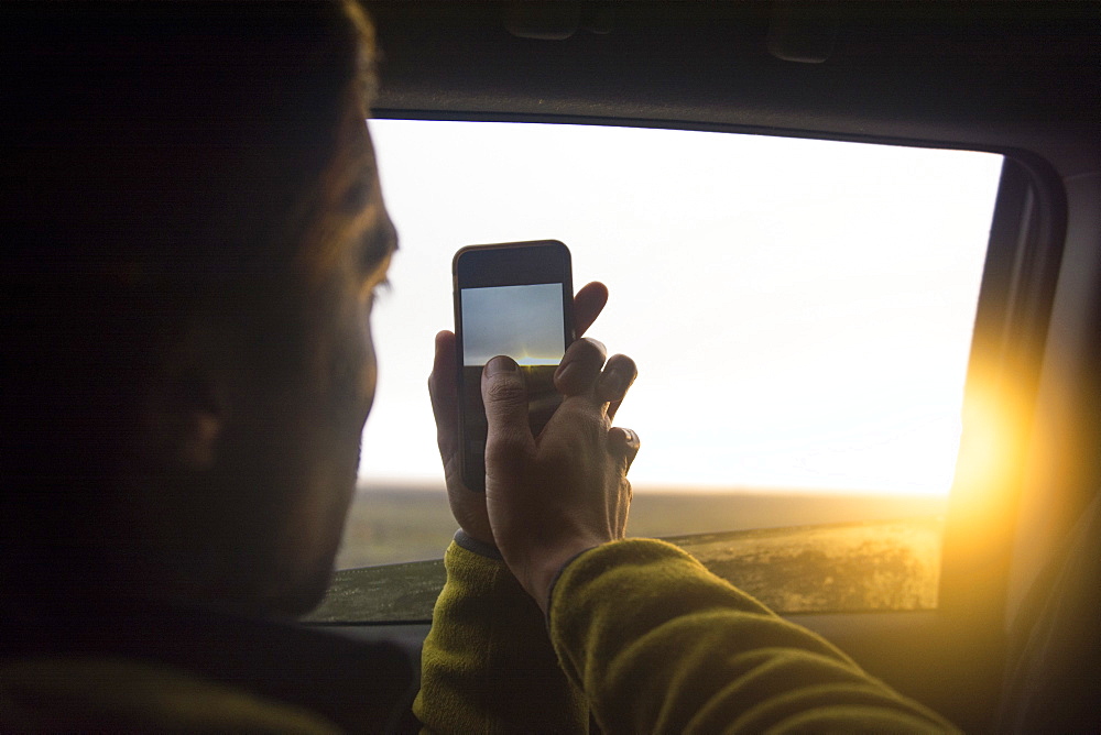 A man takes a photo of the sunset with his cellphone through the window of a car, Hofn, East Iceland, Iceland