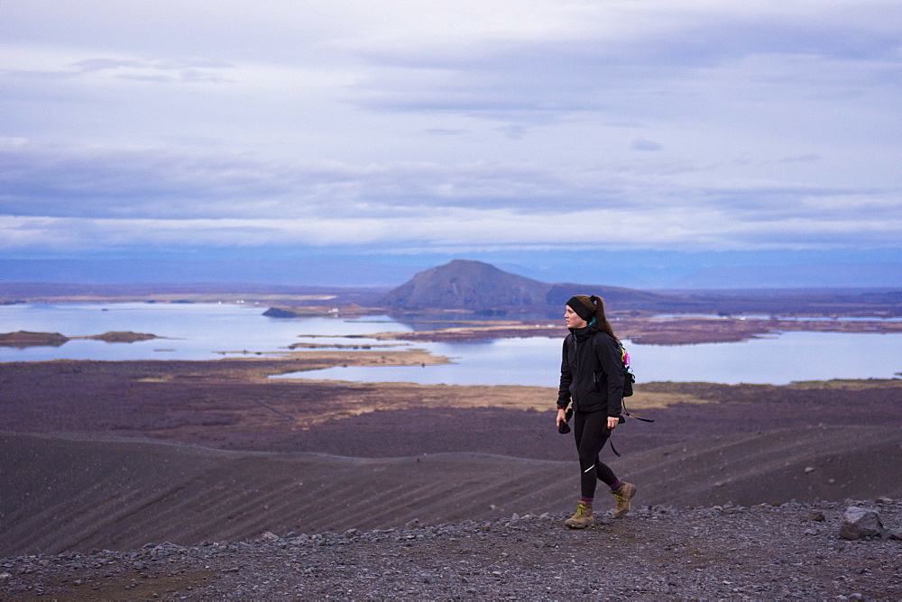 A young woman dressed in black and wearing boots walks along the ridge of the Hverfjall crater with Lake Myvatn in the background.