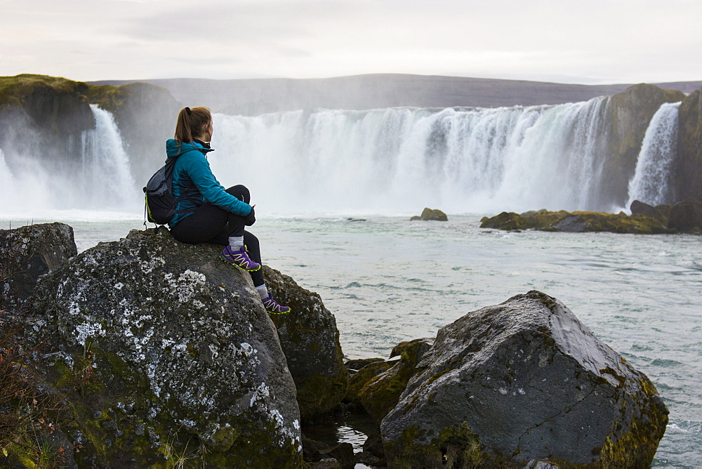 Portrait of female hiker sitting on rocks and looking at splashing Godafoss waterfall, Iceland