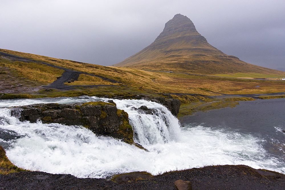 Scenic view of splashing Kirkjufellsfoss waterfall and Kirkjufell mountain on gray day, Iceland