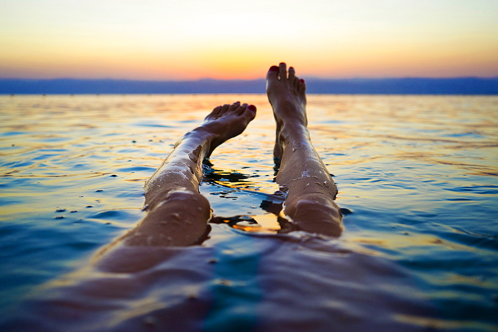 Personal perspective shot with legs of woman floating in Dead Sea at sunset, Madaba Governorate, Jordan