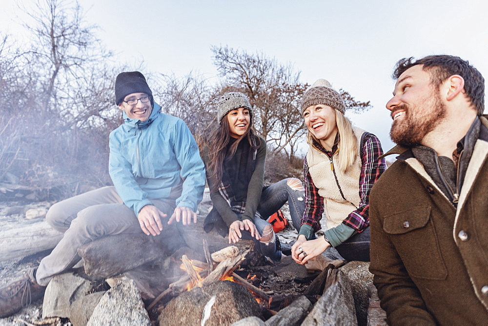 Four adult friends warming up around campfire while camping on rocky coastal beach in autumn, Portland, Maine, USA