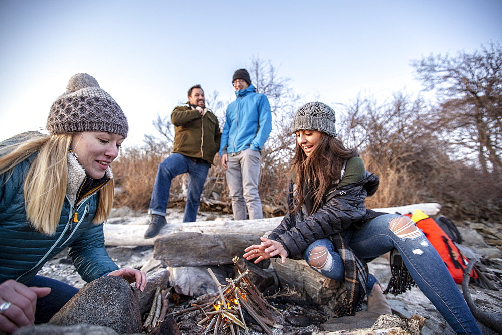 Four adult friends relaxing around campfire while camping on coastal beach in autumn, Portland, Maine, USA