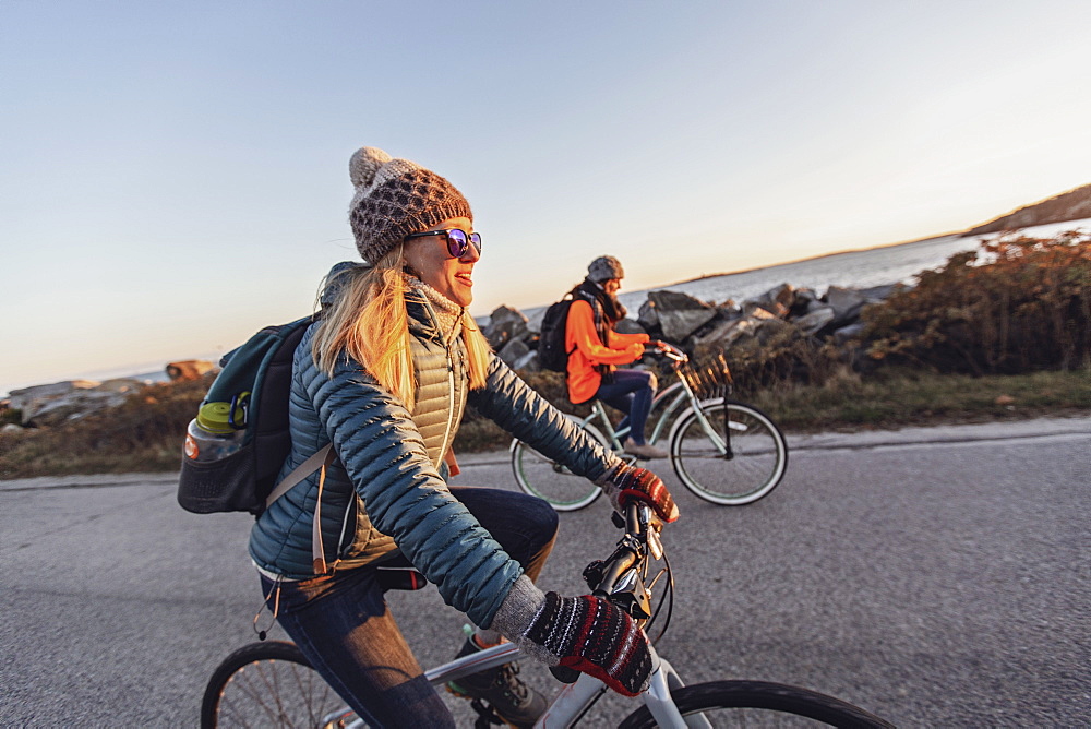 Two female friends cycling together along coastal road at dusk, Portland, Maine, USA