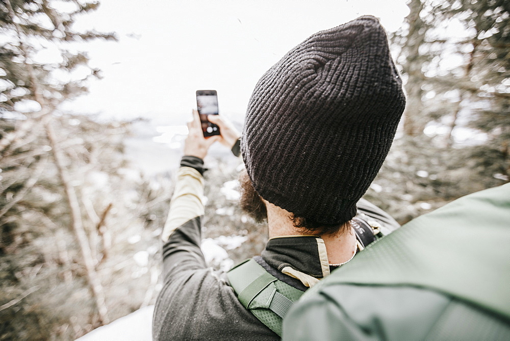 Hiker wearing wooly hat taking cell phone picture in mountain in winter, Whiteface, New Hampshire, USA