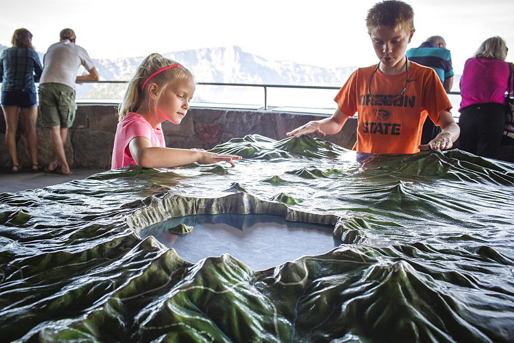 A young boy and a young girl stand at model map of Crater Lake with the real Crater Lake in the background, Oregon, USA