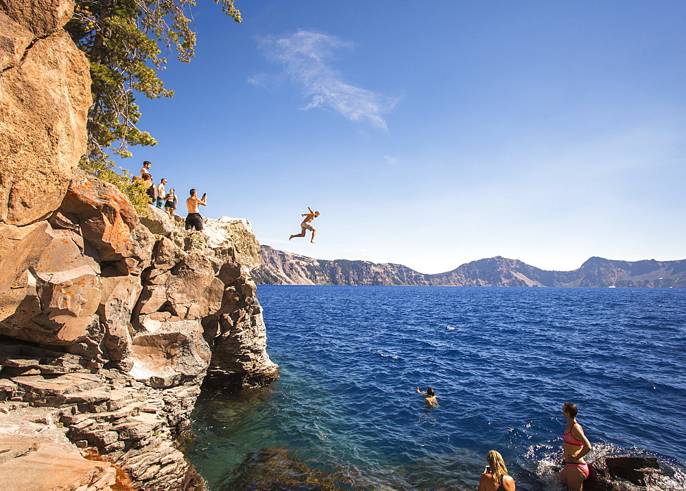 Young people wait on a rock outcrop and swim or jump into the deep blue waters of a mountain lake, Crater Lake, Oregon, USA