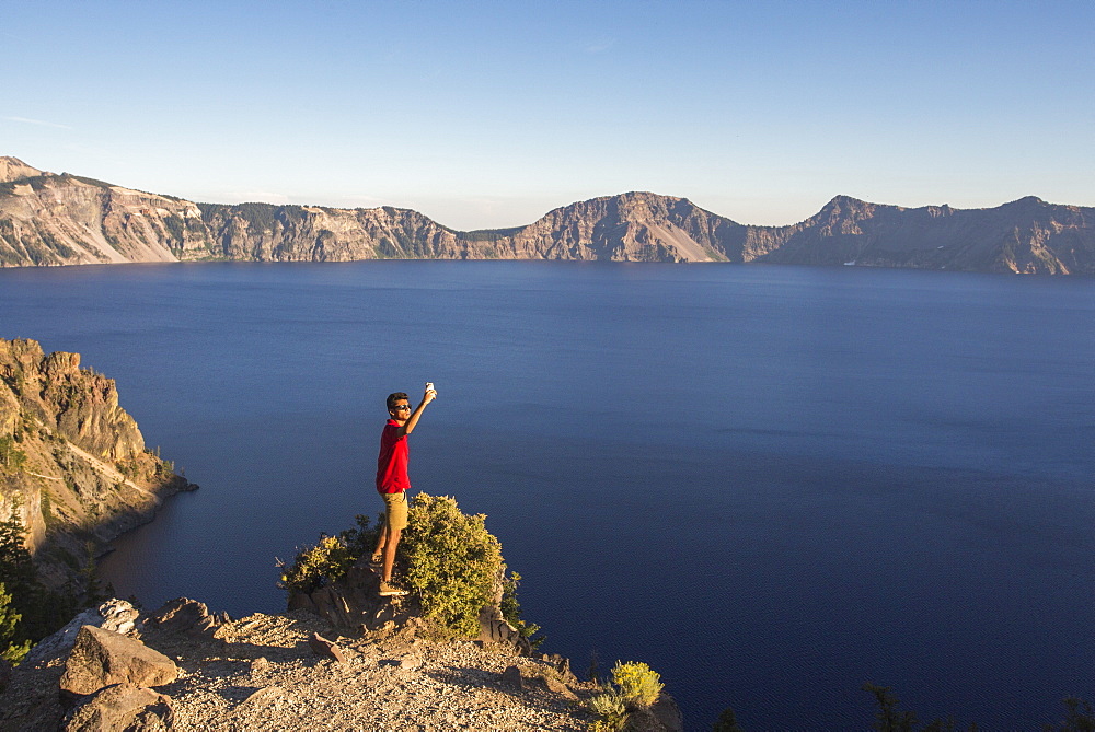 A young man in a red shirt poses for a selfie on a rock outcrop high above a deep blue lake surrounded by mountains, Crater Lake, Oregon, USA