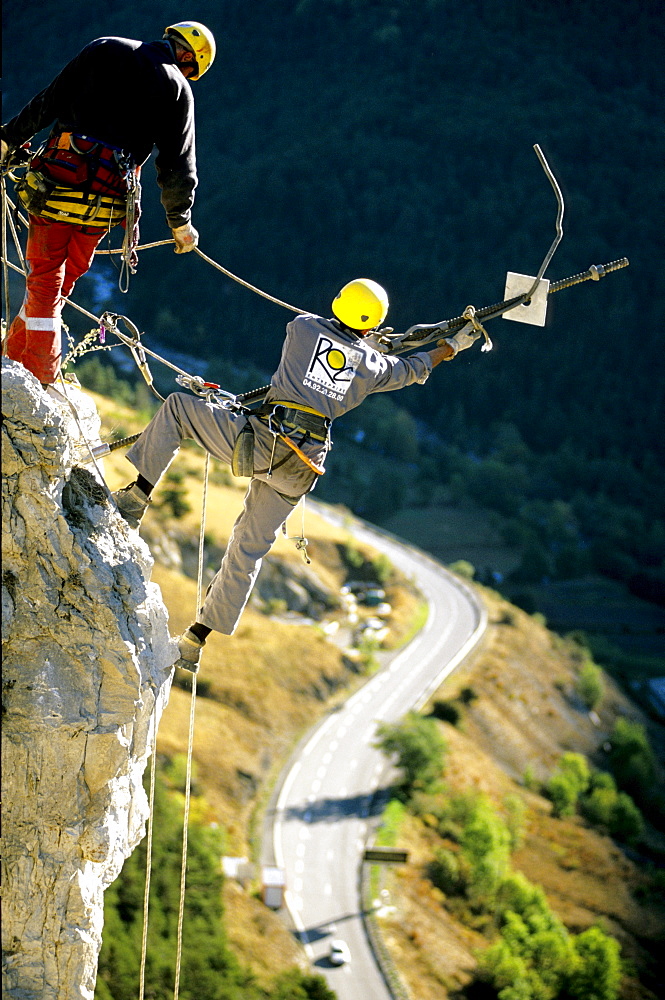 The workers, working high above the busy road below, first secure the loose boulder to the cliff with a long metal pole.