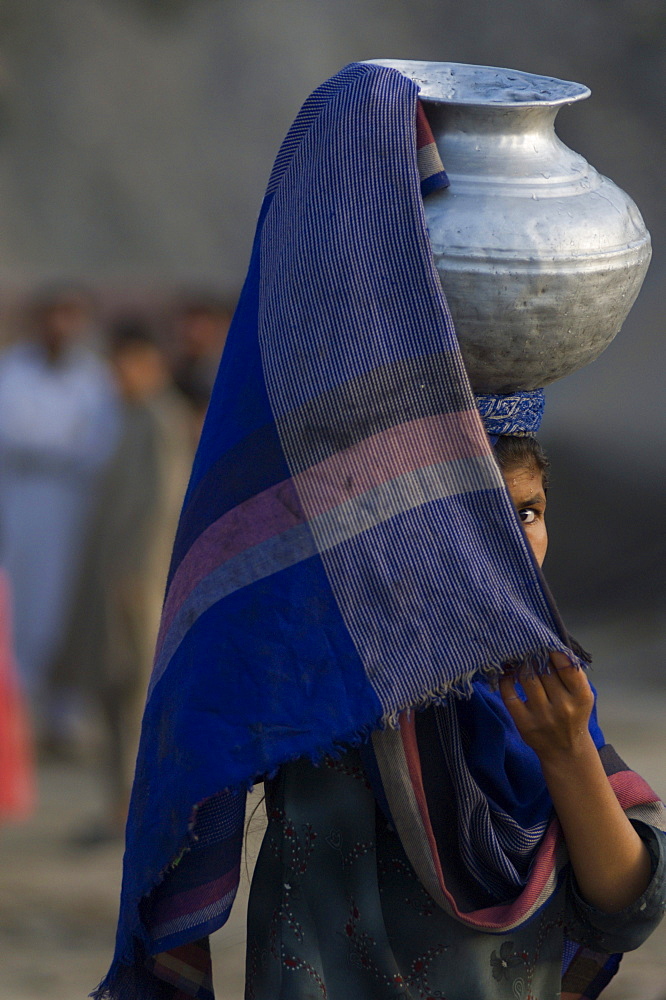 An adolescent Pashtun girl shields her face as she carries water  through the streets of the Meira camp for earthquake survivors, Northwest Frontier Province, Pakistan.  The conservative code of the Pashtun tribe of the area dictate that girls must cover themselves, including their faces, after their first period.  The crowded life in the camp has made such rules difficult to follow. The Meira Tent camp (also called Mera, or Maria camp), is located on the Indus River in the Battagram district.  The camp, the largest for displaced people in Pakistan, hosts over 21,000 earthquake survivors, primarily from the Allai valley in Pakistan's NWFP, one of the areas worst-hit by the October 8, 2005 earthquake.