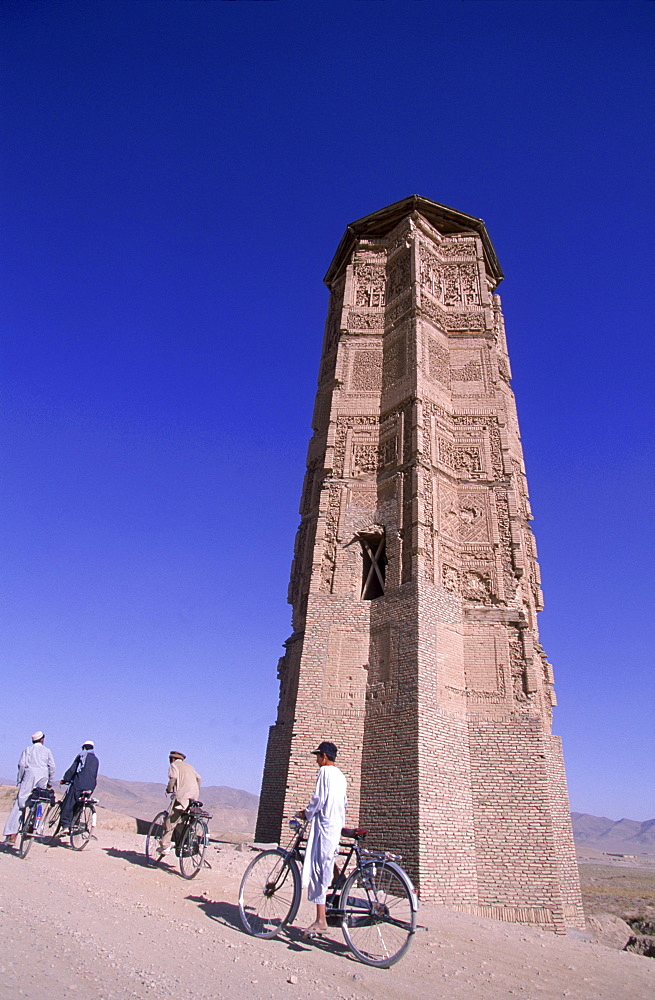 Men and boys on bikes ride past a towering, ancient  minaret, outside of Ghazni, Afghanistan, October 1, 2002. Made of brick decorated with Kufic and Naksh Script and floral motifs, the  minaret dates back to  the early 12th century and was built by Sultan Masud III of the Ghaznavid Dynasty, who ruled over an empire encompassing much of Afghanistan, Northern India, Persia and Central Asia. The minaret was once three times as tall as its current 70 feet, and is thought to have been part of a large mosque complex. Now an  important truck stop on the road to Kandahar, Ghazni, located on the Lora River at the elevation of 2,225 meters, is the capital of Ghazni province   and is a  market for sheep, wool, camel hair cloth, corn, and fruit-it also continues to be a haven for  Taliban insurgents.