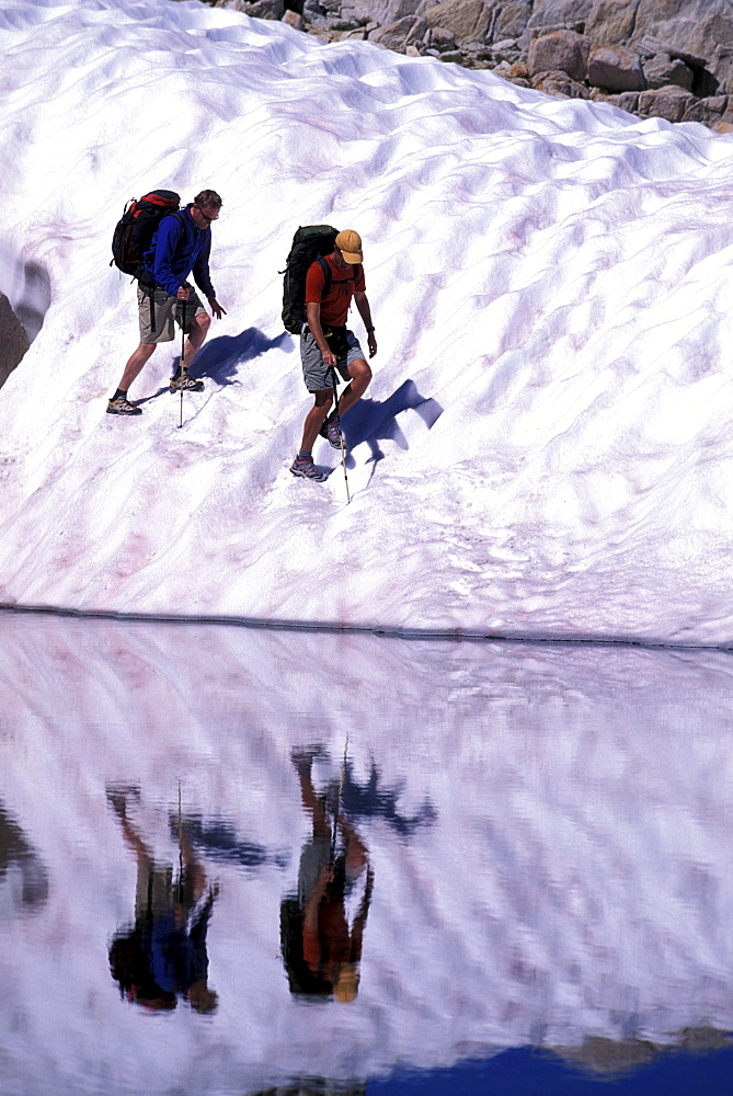 Two hikers on a glacier reflecting in a lake in Eastern Sierra Nevada mountains, California.