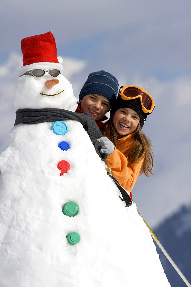 Two children peak out from behind a snowman.