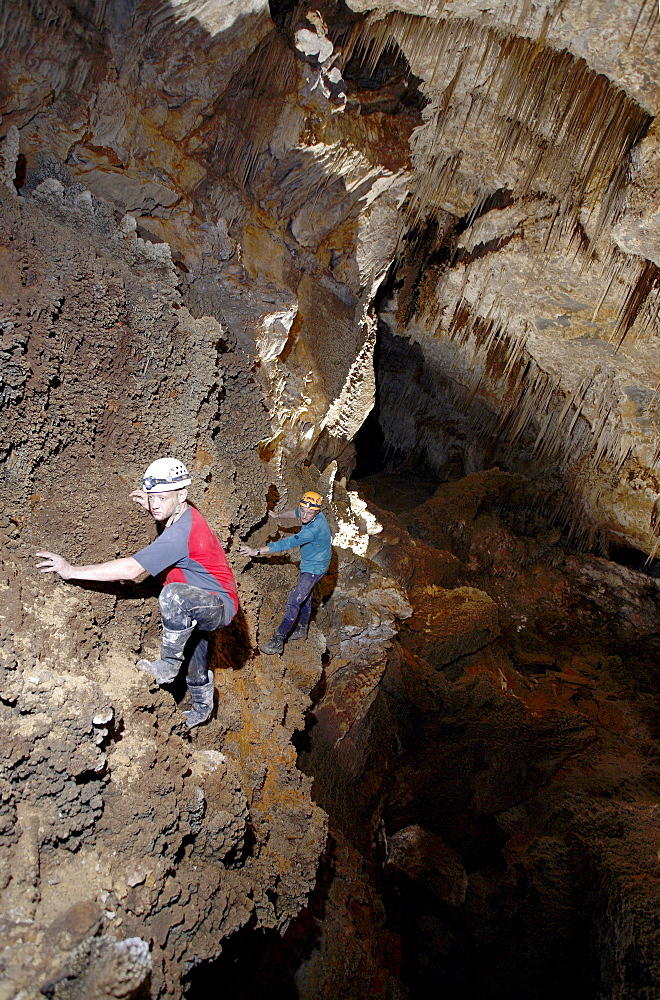 Two cave explorers traverse a section of cave on a ledge above a deep hole.