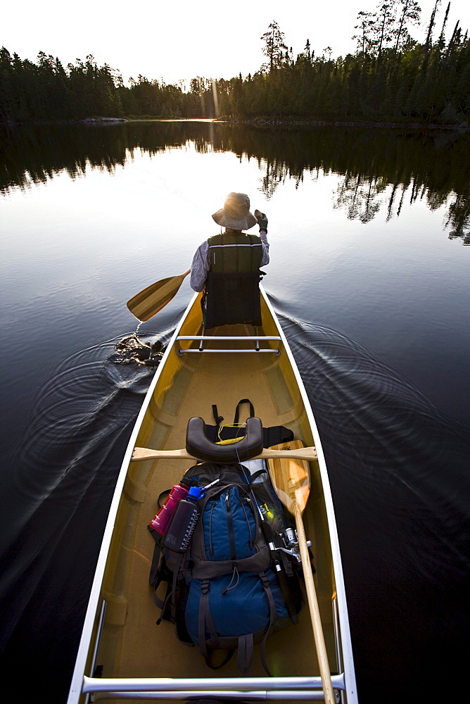 A woman paddles her canoe as the sun rises over the trees on the horizon of the Boundary Waters in Minnesota.