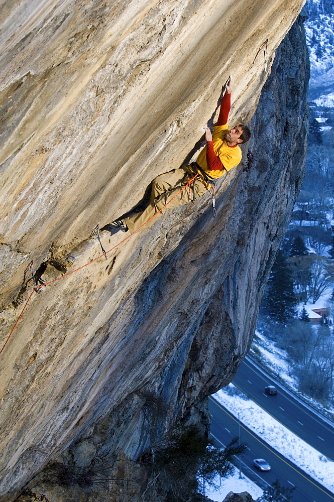 A rock climber in a yellow and red shirt reaching up for a hand hold on a steep and difficult route.