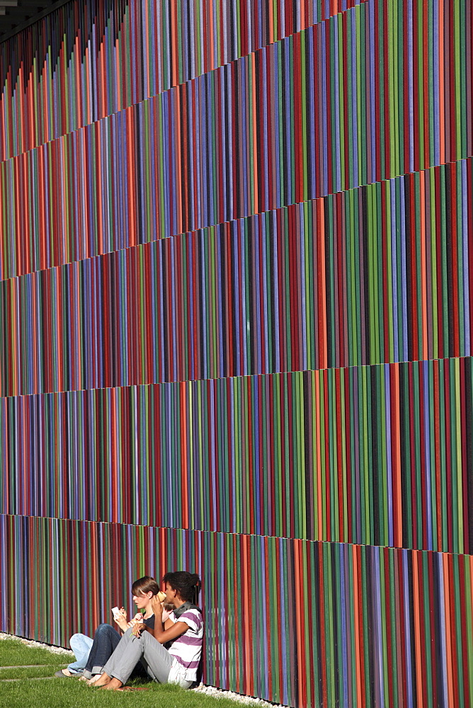 Two girls sit against a colorful wall outside the Brandhorst Museum in Munich, Bavaria, Germany.