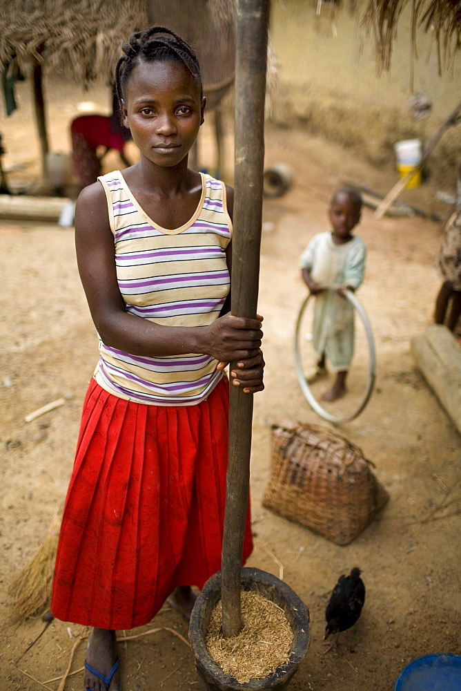 Young mother working outside her home with her young son in the background playing with an old bike rim. Gbolokai is a small town of a few hundred people 20 minutes off the main road near Totota, all of its inhabitants fled during the long brutal civil war and have slowly returned after 2005 to try and rebuild their lives.