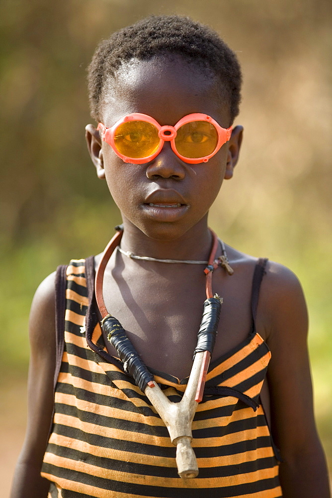 Portrait of a young boy wearing bright orange glasses and a sling shot around his neck. Gbolokai is a small town of a few hundred people 20 minutes off the main road near Totota, all of its inhabitants fled during the long brutal civil war and have slowly returned after 2005 to try and rebuild their lives.