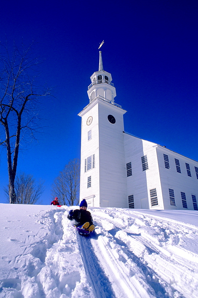 A boy sledding on a snowy hillside in front of the church in Strafford, Vermont on a clear, cold winter day.