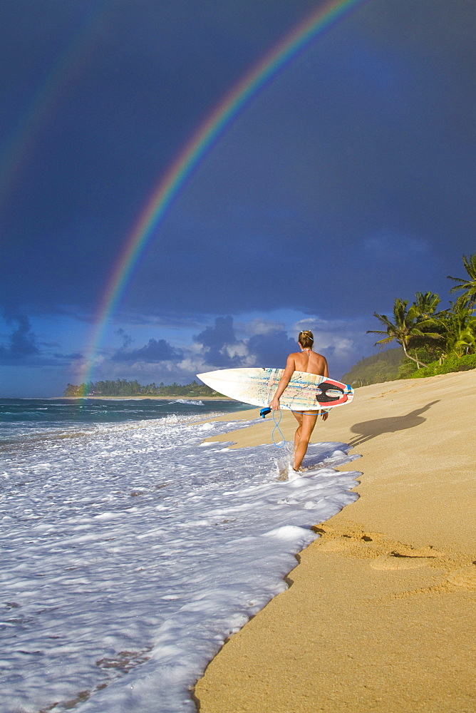 An amazing rainbow over Rocky Point, as a surfer girl walks along the beach, on the north shore of Oahu, Hawaii.