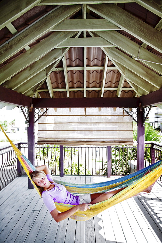 A girl relaxes on a porch in a hammock  with her arm behind her head.