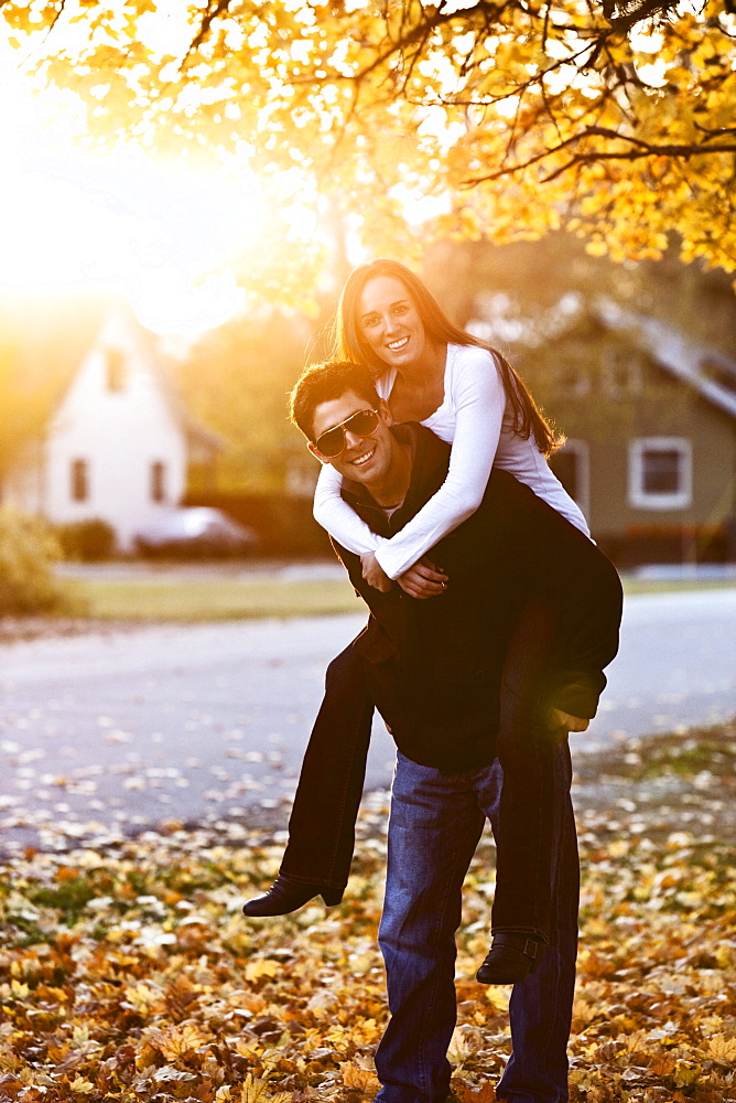 A young adult couple smiling while piggy back riding at sunset surrounded by fall colors in Sandpoint, Idaho.